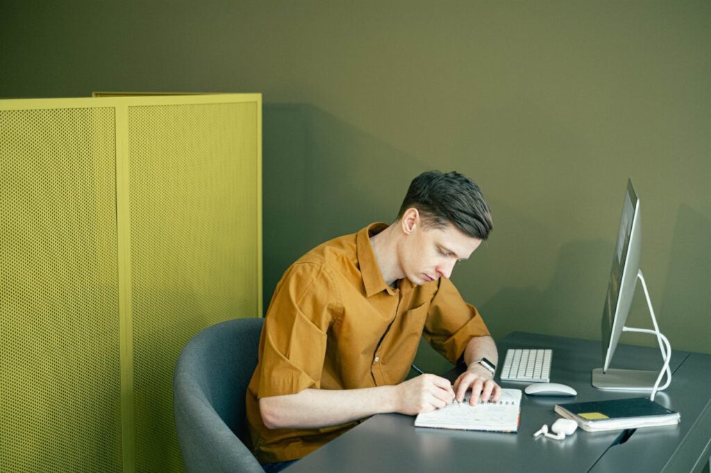 a man in a yellow shirt sitting at a desk sketching logos with a monitor in front of him, looks like an apple computer. A yellow screen is also behind him.