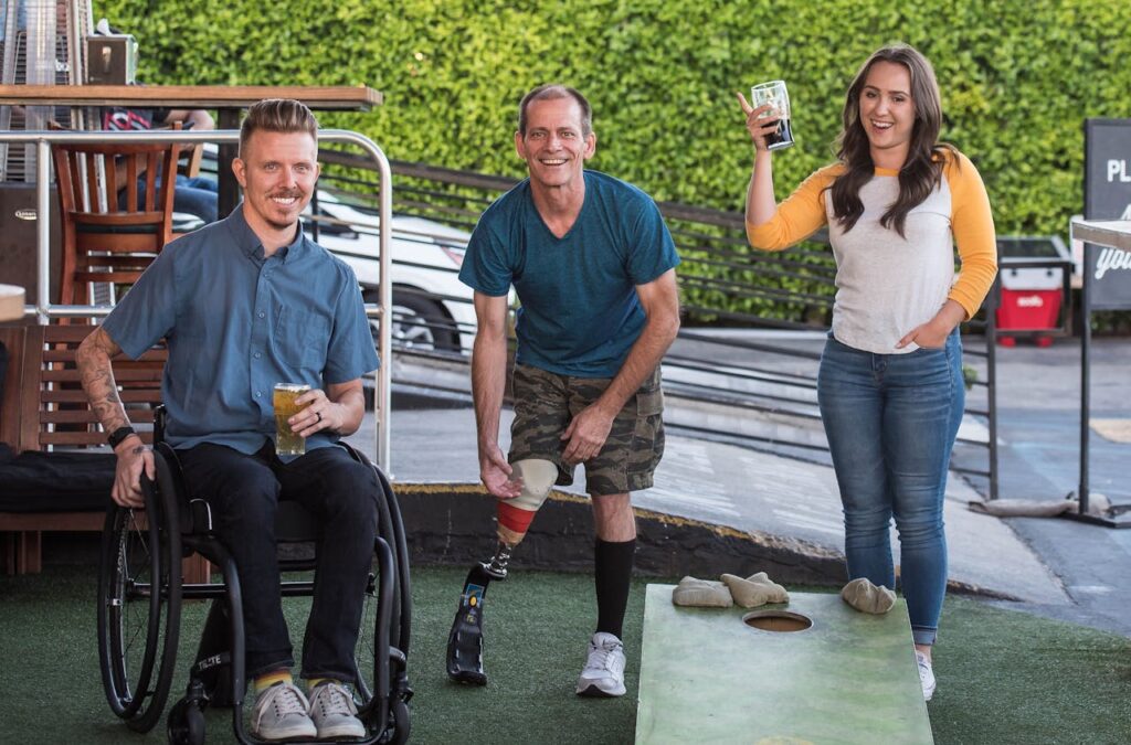 a young man in a wheelchair holding a pint of beer. A middle aged man with a false leg and a lady, about 30 years old holding what looks like a glass of coke. All smiling directly at the camera. This is to represent the importance of designing for all in 2025