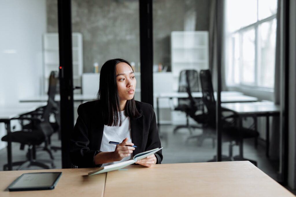 an asian woman wearing a suit, with a relaxed tshirt underneath, thinking, with a book in her hand, looks like she is in an office, she could well be a branding consultant or a business owner thinking about her brand